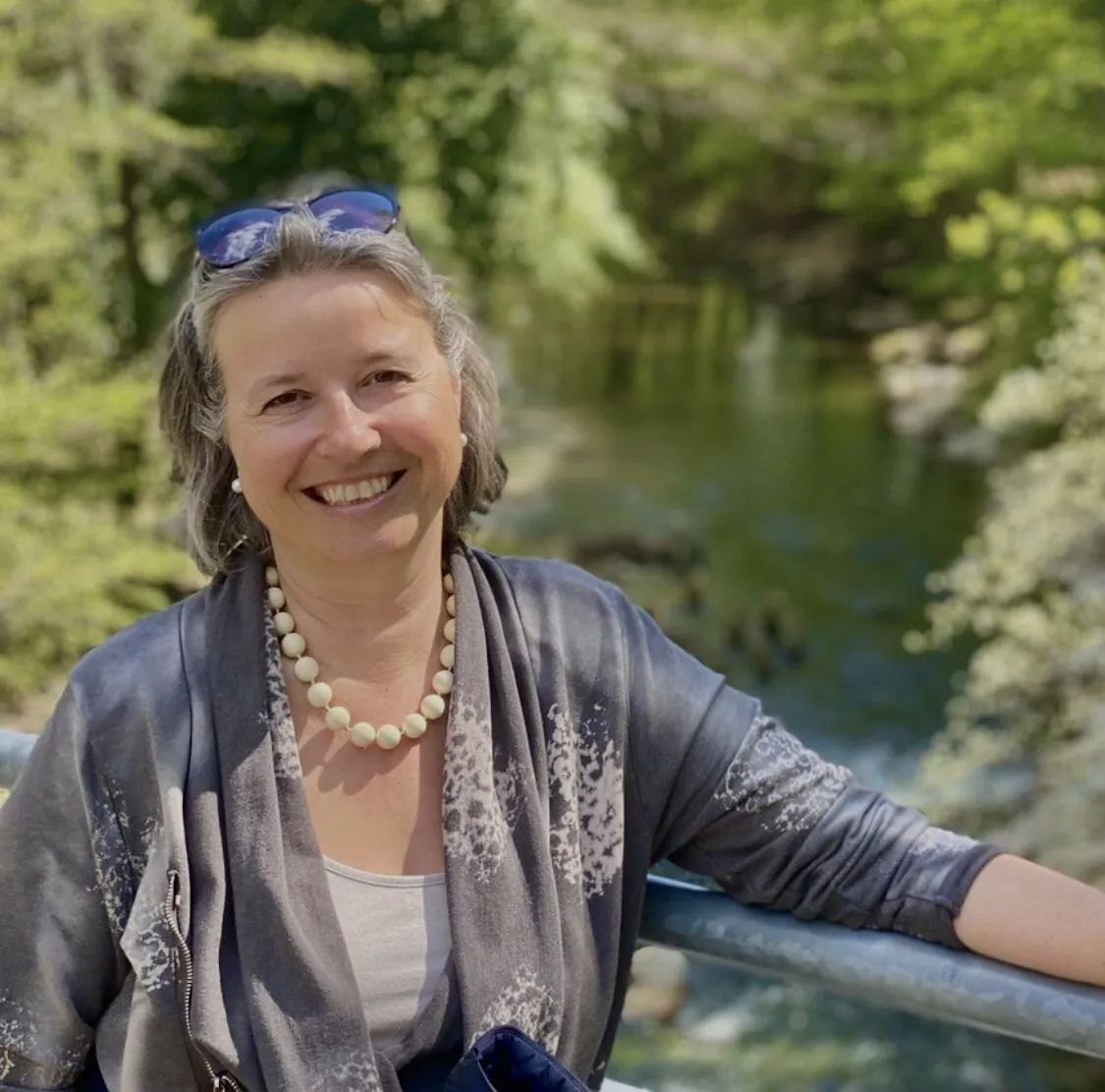 Karin Daurer lächelt, an ein Geländer gelehnt, vor einem Bach im grünen Wald